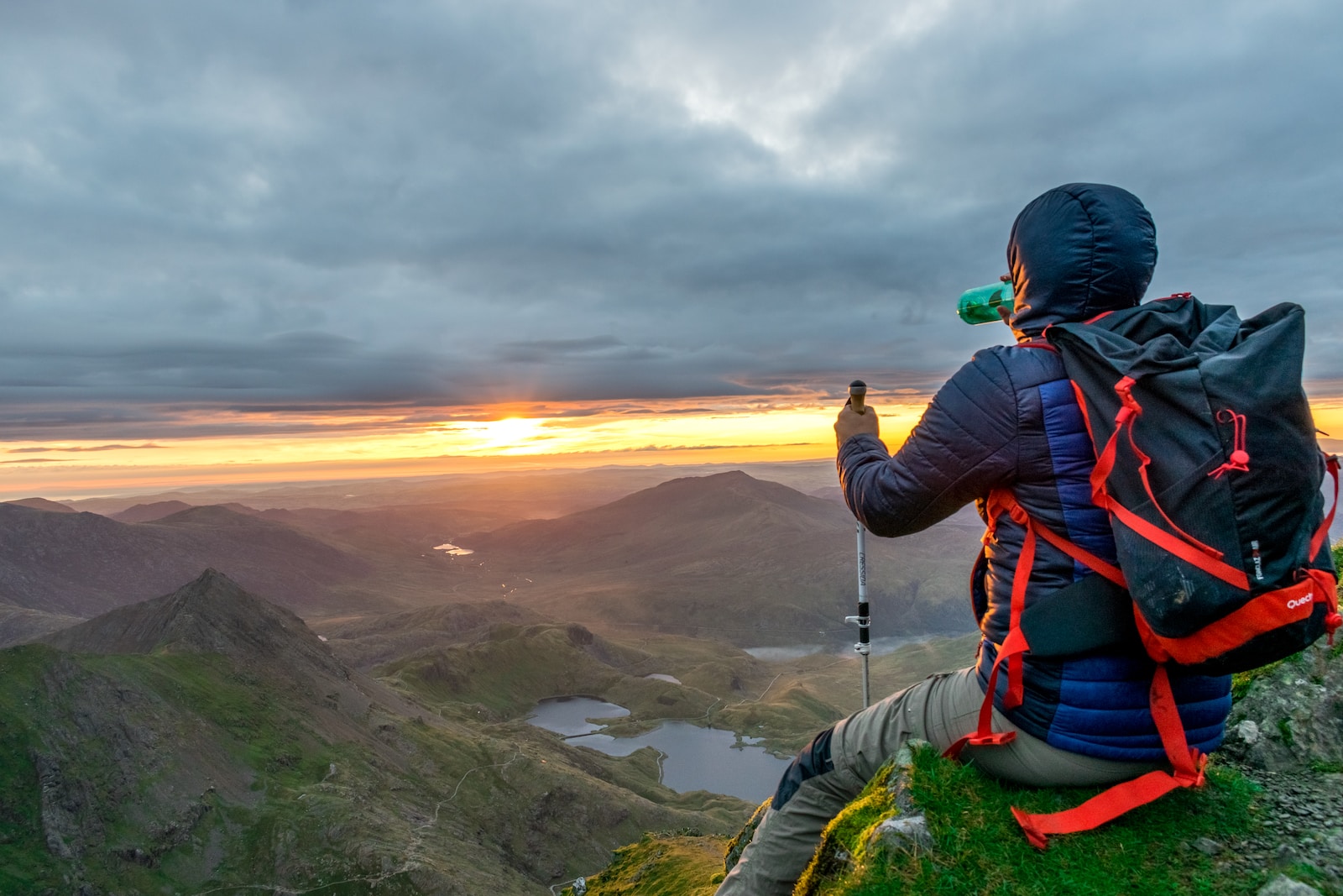 man sitting on cliffhanger looking at mountain under gloomy clouds